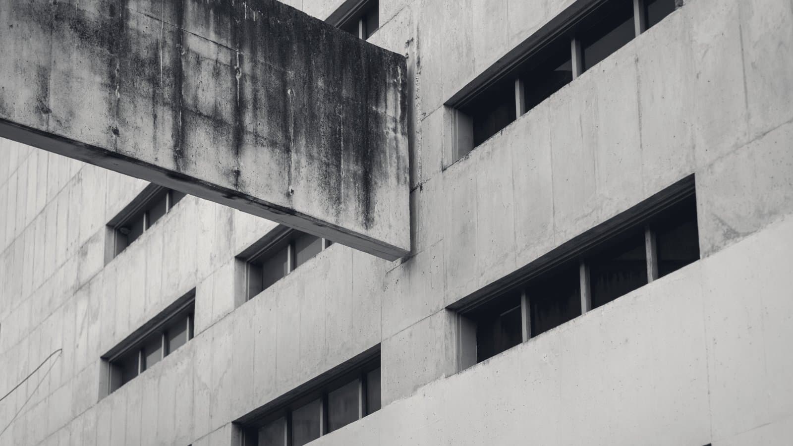 A heavy cantilevered concrete slab projecting across the face of a windowed concrete building, blocking a whole run of openings behind it.