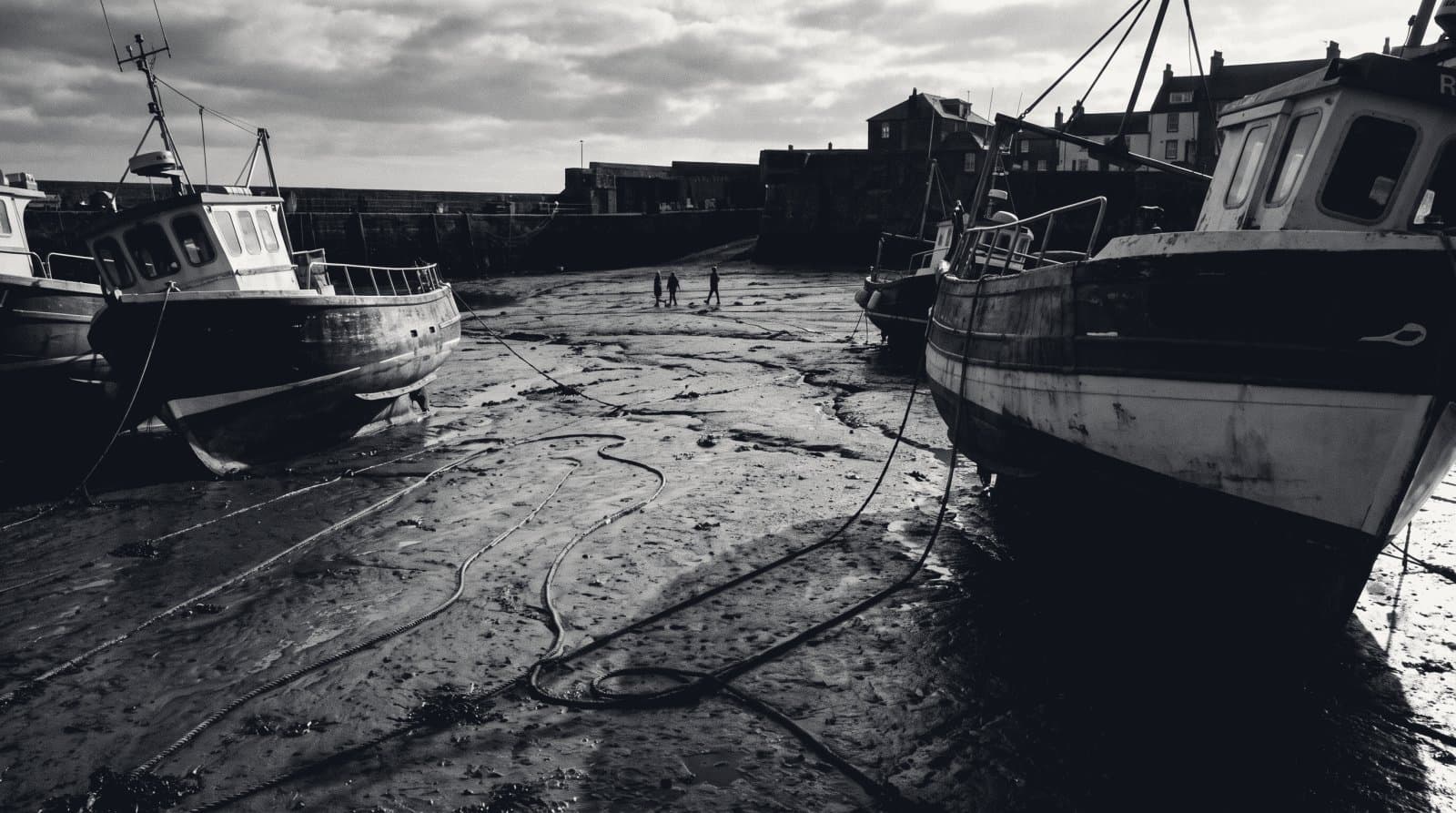 Fishing boat hulls resting on harbour mud at low tide, mooring lines slack.