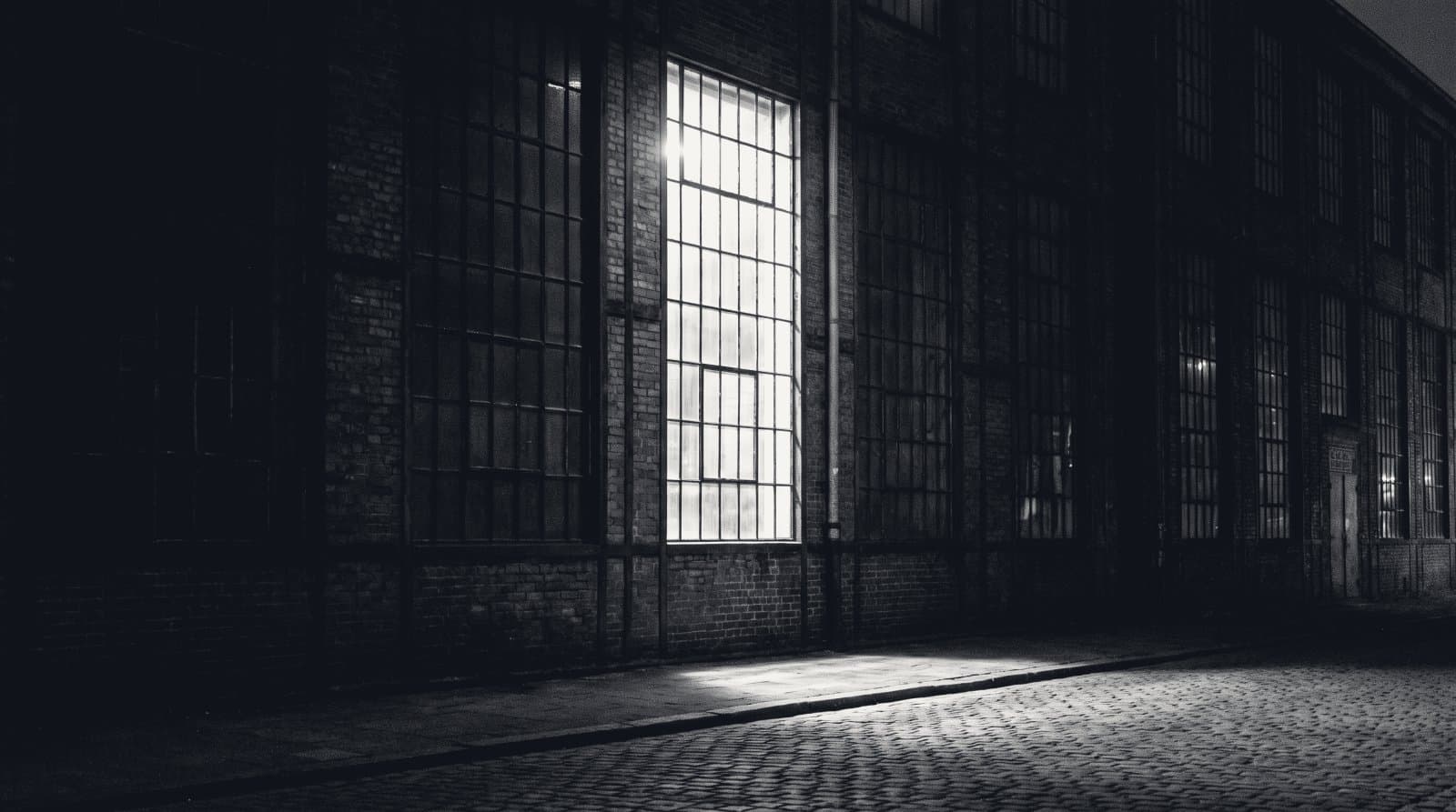 The steel-framed window wall of a small brick industrial workshop at night, one lit bay among dark ones.